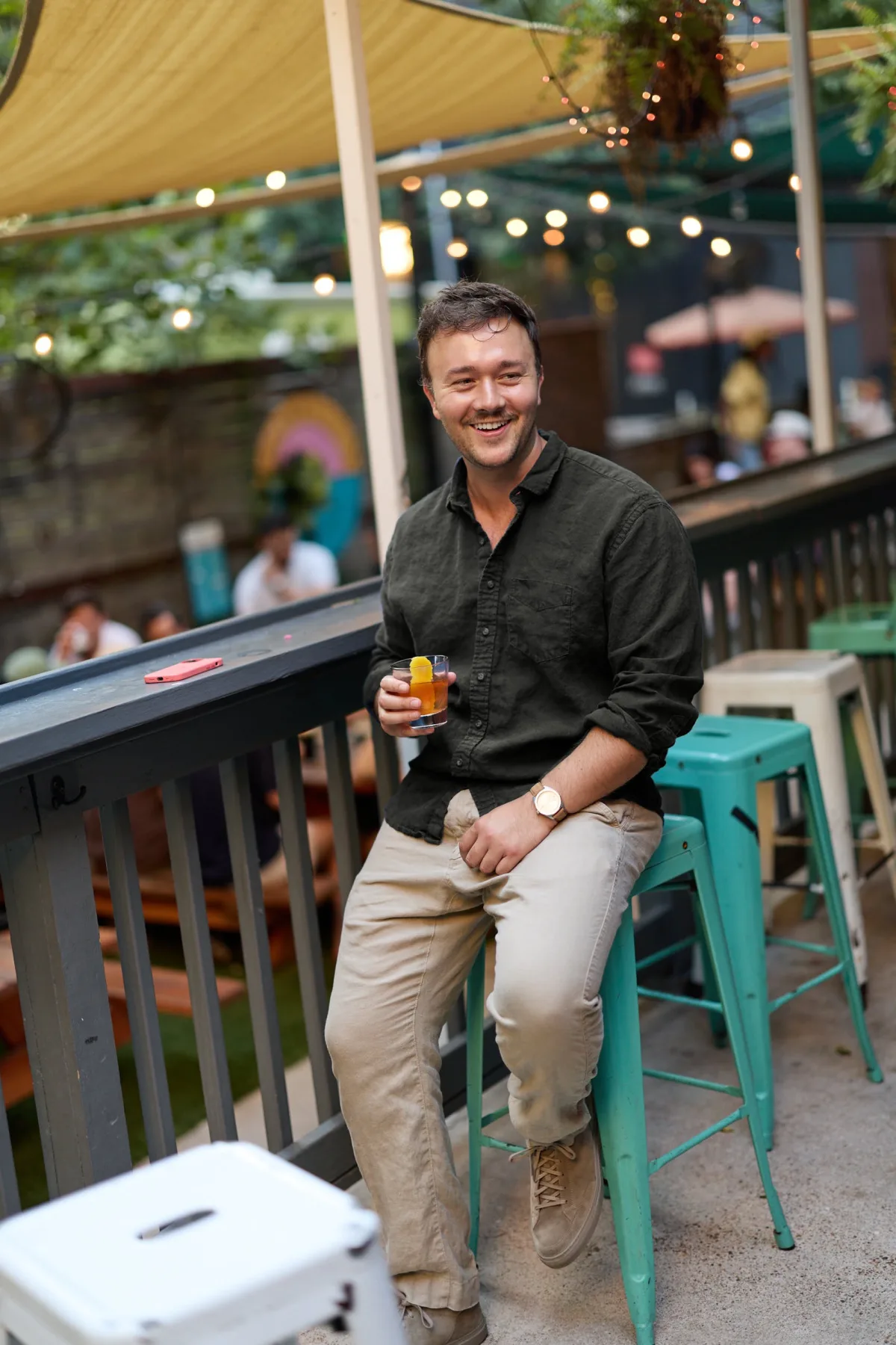 Man with a drink on a bar patio stool under string lights