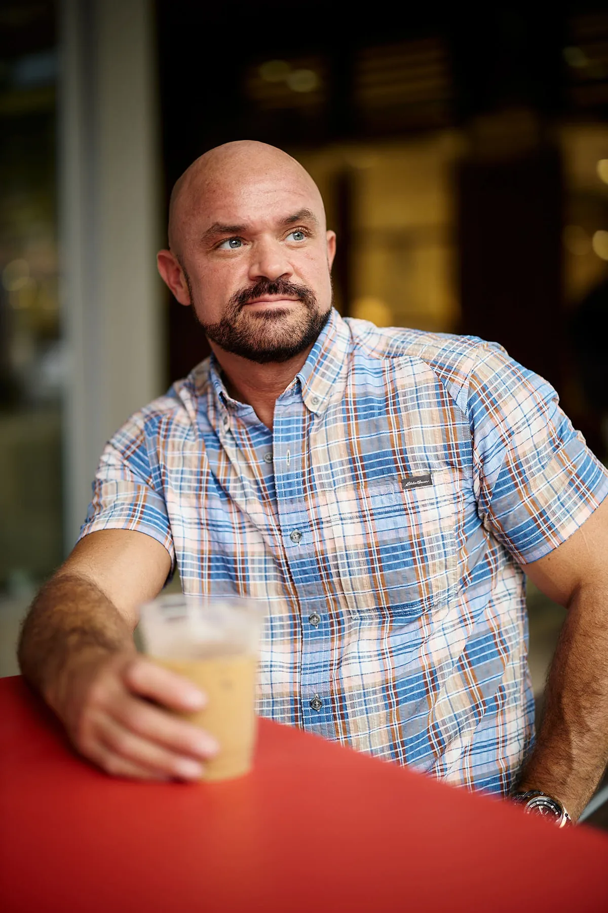 Man in a plaid shirt with a drink at a red table, a relaxed dating profile portrait