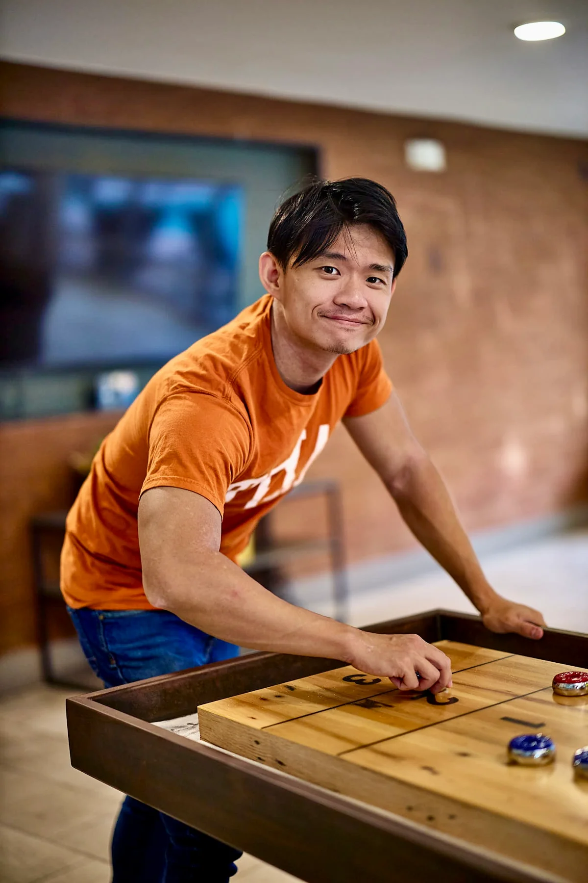 Man in an orange Texas tee mid-move at a shuffleboard table, a candid reaction shot for a dating profile