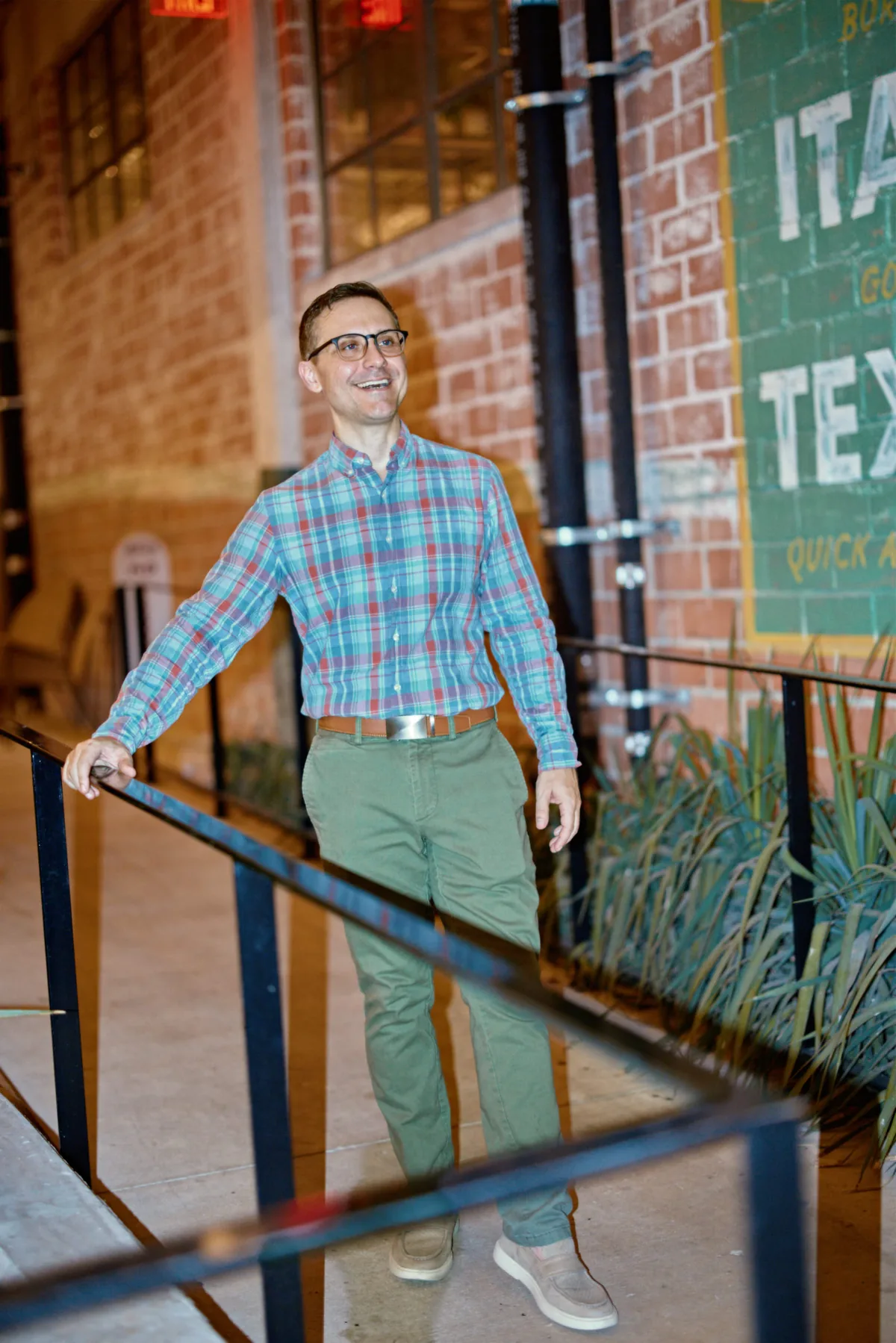 Man in a plaid shirt and green chinos laughing by a brick wall, a full-body style shot for a dating profile