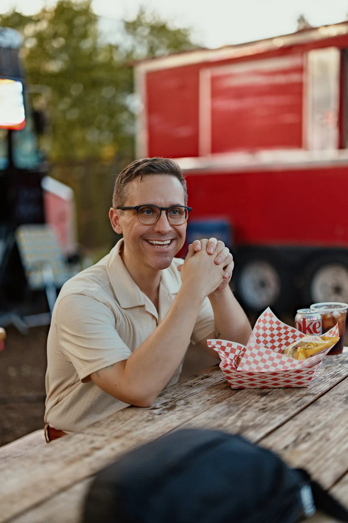 Man laughing at a picnic table beside a red food truck with a basket of food, a candid dating profile photo