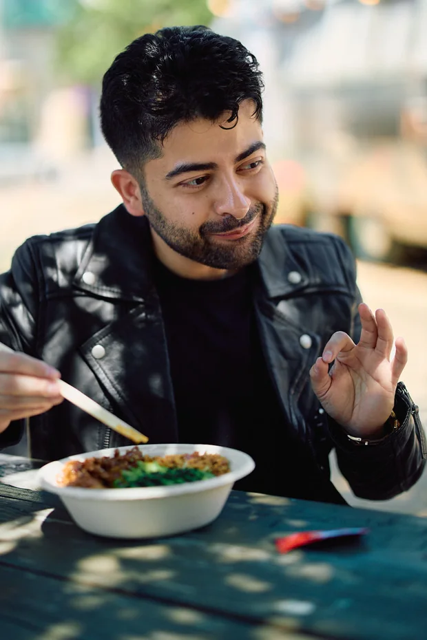 Man in a leather jacket eating with chopsticks at a food truck patio, giving an okay sign, a candid dating profile photo
