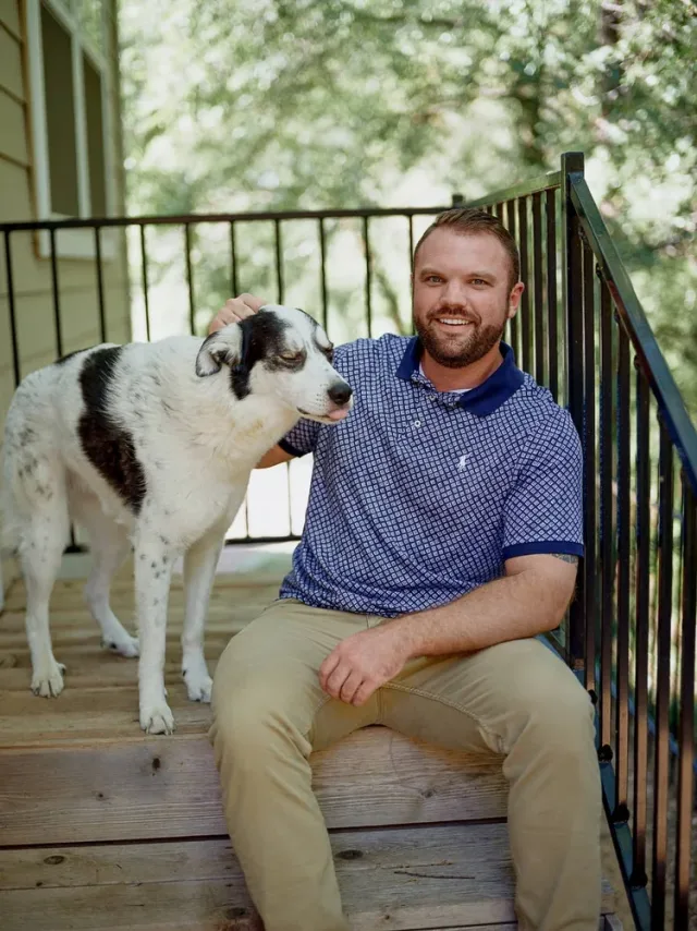 Portrait of a man sitting on wooden steps beside a black and white dog, with leafy trees and soft natural light behind him