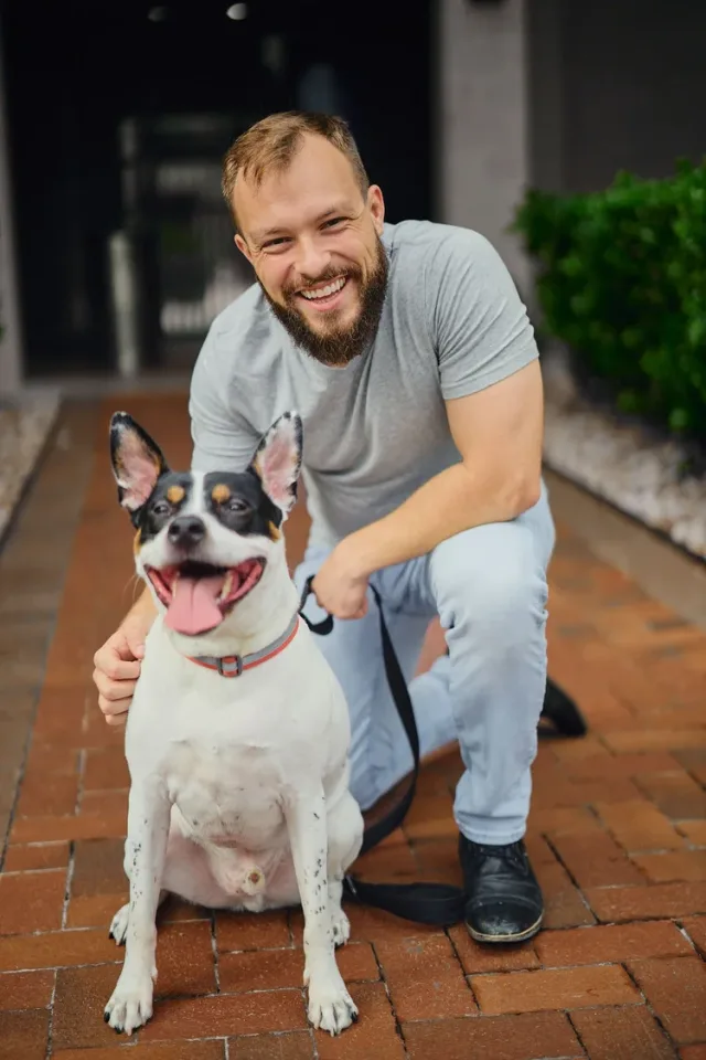 Smiling bearded man in a grey t-shirt crouching beside a happy black and white dog on a brick sidewalk