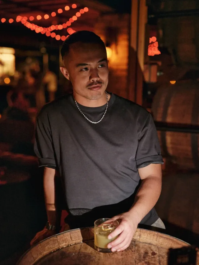 Portrait of a man in a dimly lit bar holding a cocktail beside a wooden barrel, with warm string lights behind him