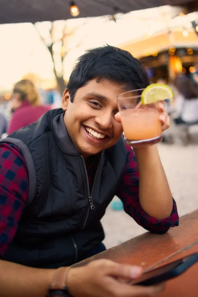 Man holding a drink at an outdoor bar at sunset, smiling at the camera in a candid Austin dating profile moment