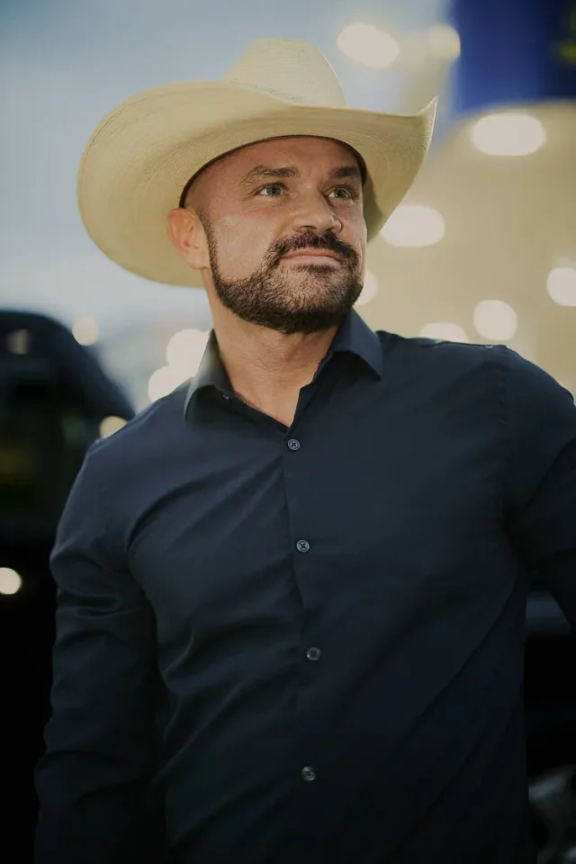 Man with a beard wearing a light cowboy hat and dark shirt, with out of focus lights behind him, a hero shot for a dating profile