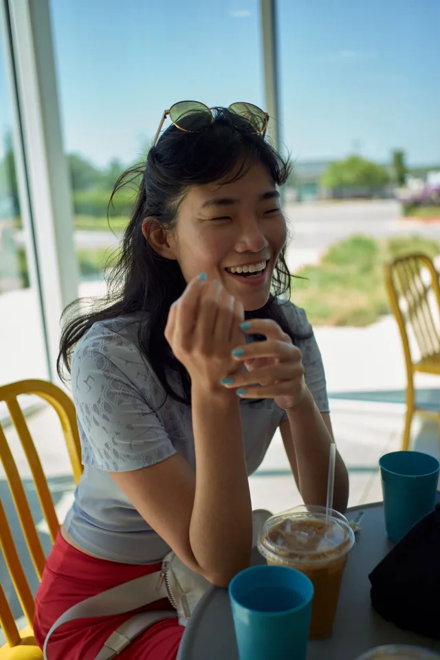 Woman laughing at a cafe table with an iced coffee and sunglasses on her head, a candid dating profile photo